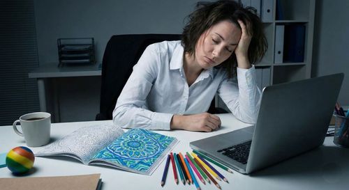 An exhausted professional sitting at a desk, eyes closed for a moment, next to an intricate half-colored illustration and a set of professional pens.