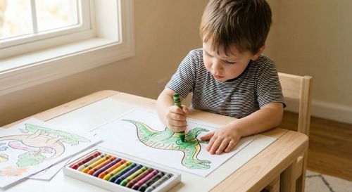 A young child sitting at a colorful desk, deeply focused on coloring a simple animal drawing with a bright green crayon.